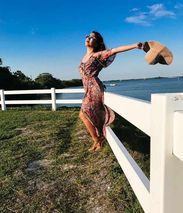 Girl in pink flowery dress and straw hat leaning against a white fence by the sea, holding her hat behind her head on a bright day - Smartphone fashion photography shoot
