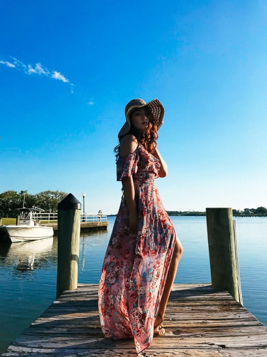 Girl in a long flowery dress and straw-hat standing on a wooden dock on a bright day, blue water and skies in the background. Smartphone fashion photography