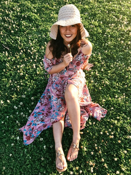 Girl in pink flowery dress and straw hat sitting on the grass and holding a white flower under her chin on a bright day - Smartphone fashion photography shoot