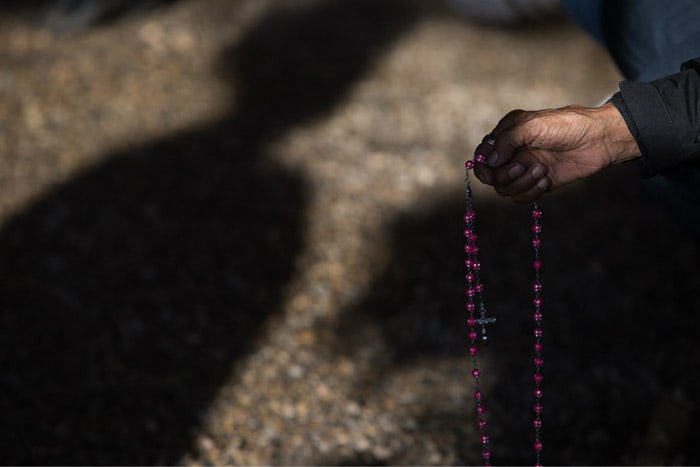 a hand holding rosary beads against a shadowing background. Photo by Amber Bracken.