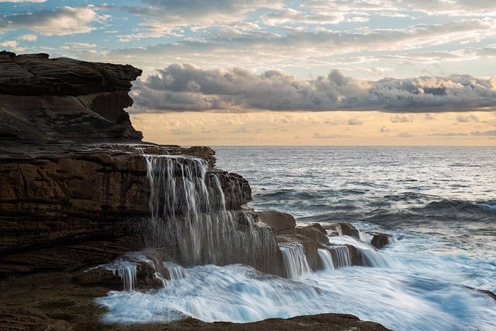 Seascape photo of a waterfall running into the ocean, cloudy skies.