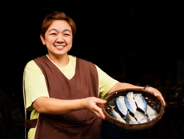 A smiling woman poses with a basket of fish for portrait against a dark background