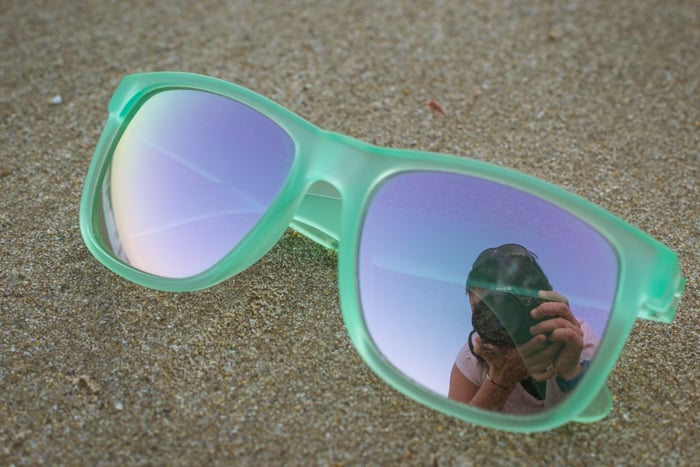 A pair of green framed sunglasses resting on the sand with the photographers reflection in one glass, cool beach photography ideas. 