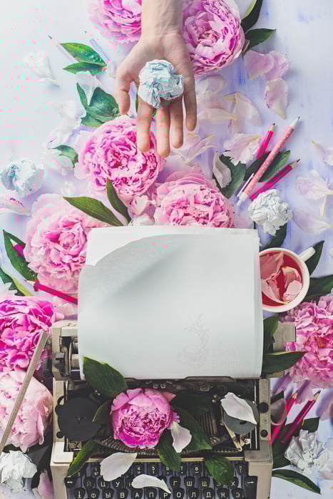 A bright and airy flat lay still life with a typewriter, pink roses and a hand holding a crumpled ball of paper