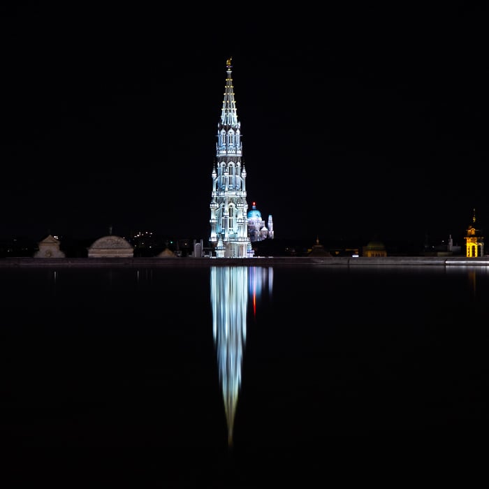A reflection on the fountain in Mons des Arts against a black night sky. Town hall, Grand Palace, and Basilica of the Sacred Heart in Brussels in the background.