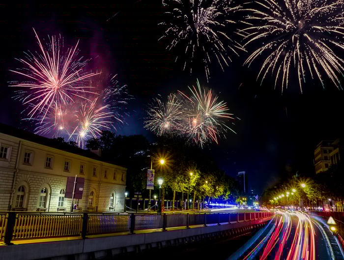 an urban street and building lit by colorful fireworks and light trails from vehicles on the street against a dark night sky