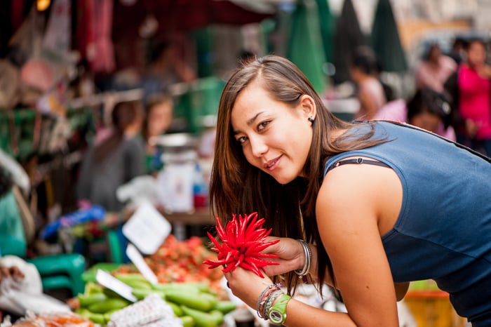 A brunette woman in a sleeveless navy shirt smiling and leaning over wares in a fresh marketplace