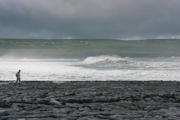 A lone silhouette of a man walking along a beach with big waves behind him.