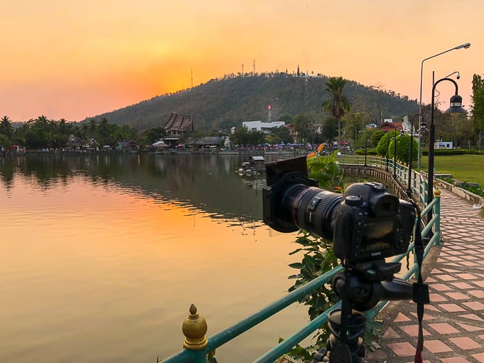 camera set up on a tripod at the shore, looking over at the mountain, sea, and horizon in the golden sunset - How to Take Stock Photos That Sell