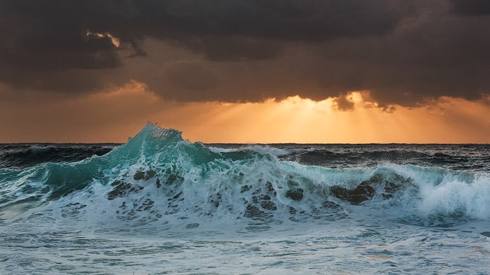 foamy waves crashing onto the shore, golden sun beams peeking from the dark clouds on the horizon. Shutter speed is 1/50 sec