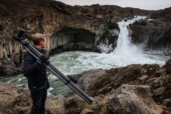 A person standing in front of a large waterfall, holding a camera and standing on rocks with ice formations around them.