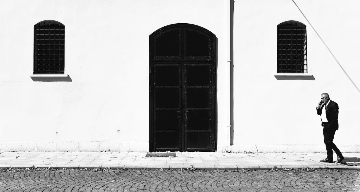 Black-and-white photo of a man in a suit walking on a sidewalk with a building facade backdrop