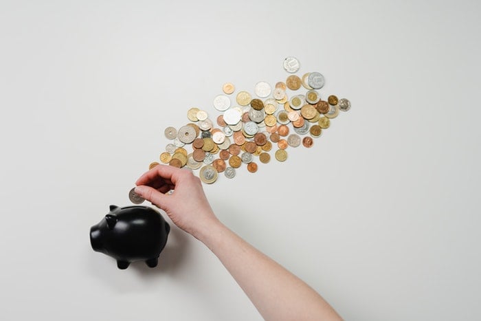 A hand holding a piggy bank filled with coins while a stream of coins flows out of the top.
