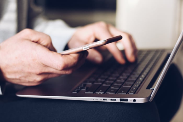 Closeup of a man's hands while typing on a laptop and holding his mobile in his other hand.