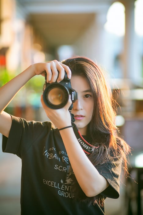 A woman holding a camera and taking a photo, standing outdoors with blurred background.