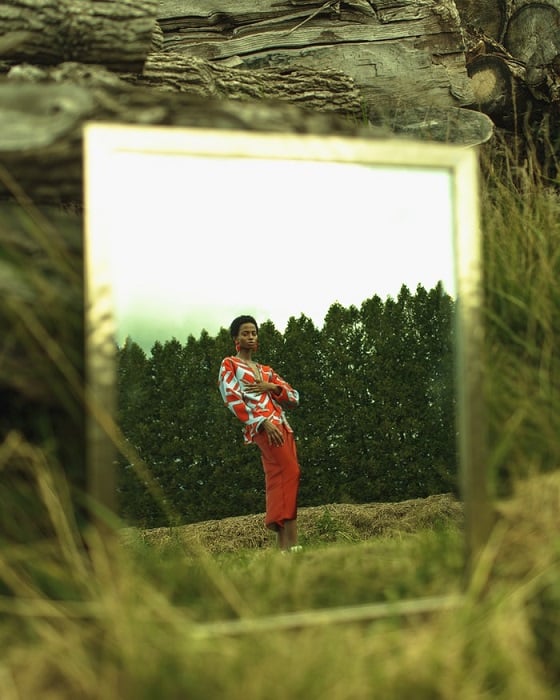Editorial photo of a mirror with a reflection of a woman standing in nature with trees in the background