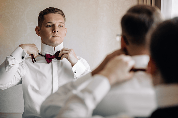 groomsmen photo idea: Groom in mirror having his bowtie adjusted by a groomsman