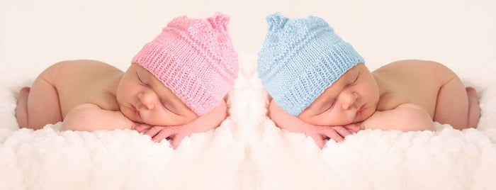 newborn twins photography: newborn twins in pink and blue knitted hats posed with their heads resting on their crossed arms