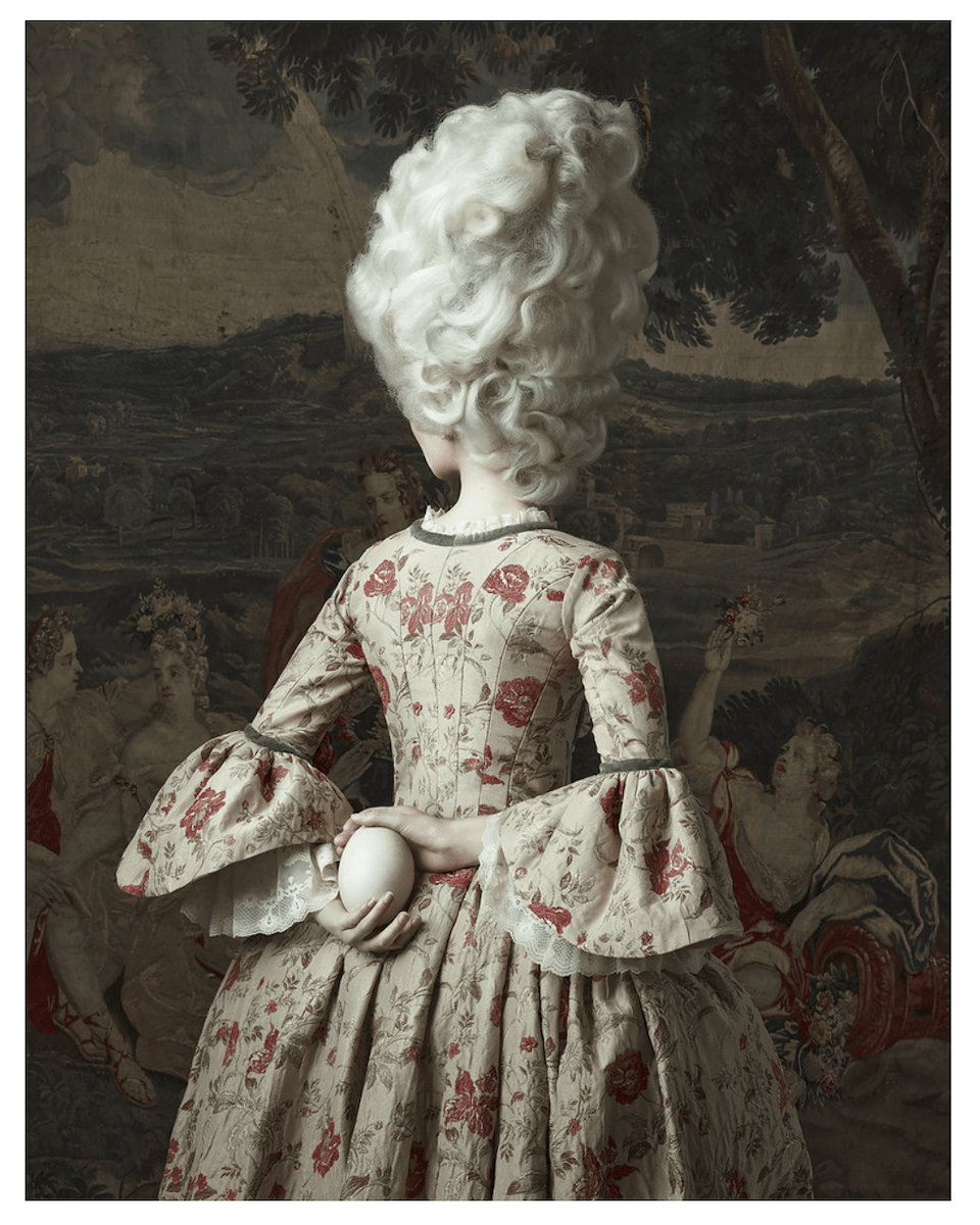 A woman dressed in Victorian-style with her back turned and holding an egg behind her back