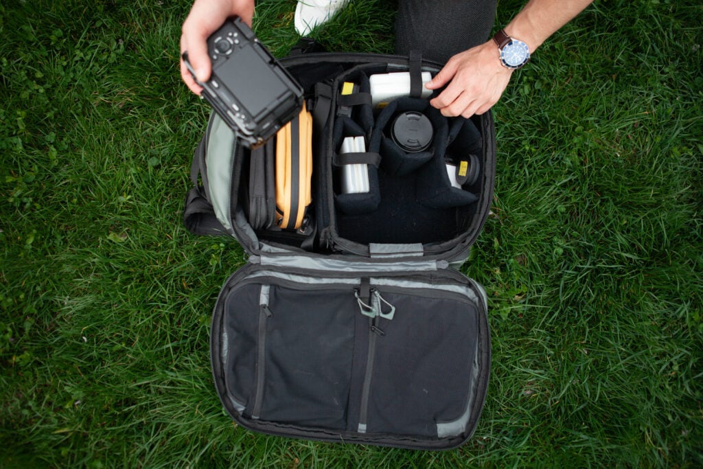 An open black camera bag with various items inside, being shown by a person standing in the background.