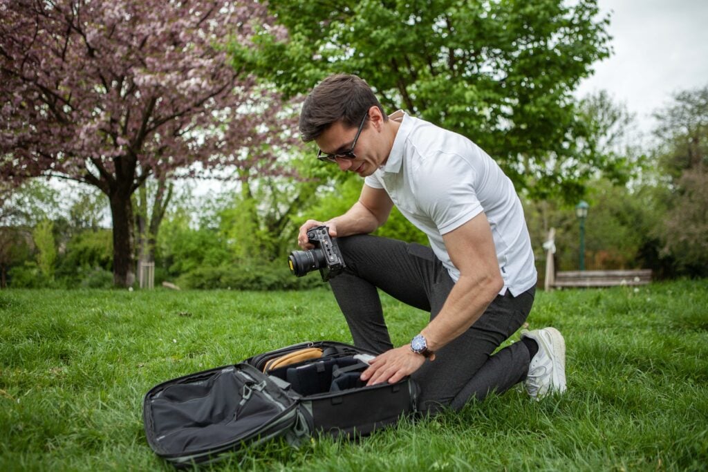 A man in a white shirt is kneeling on the grass, next to his luggage.