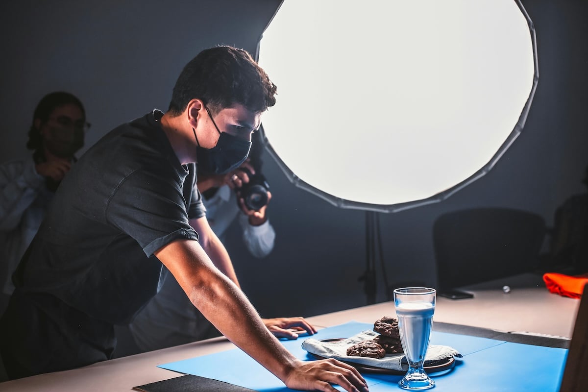 In a dark room, a man in glasses takes a photograph of someone sitting at a table with chocolate chips cookies.