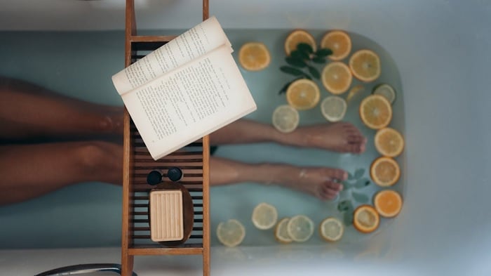 Overhead shot of woman's legs in a bathtub with fruits slices in and a book on a over-bath shelf