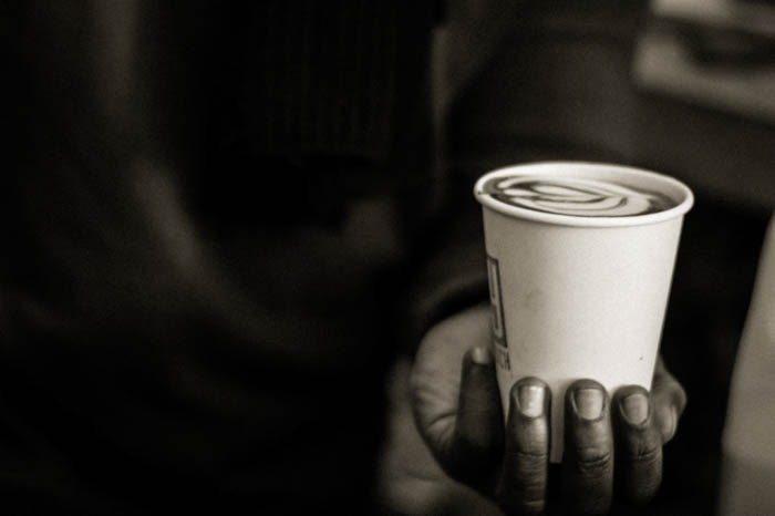 Närbildsfoto av en persons hand som håller en vit kaffemugg i plast. Den kreativa latte-designen är synlig. 