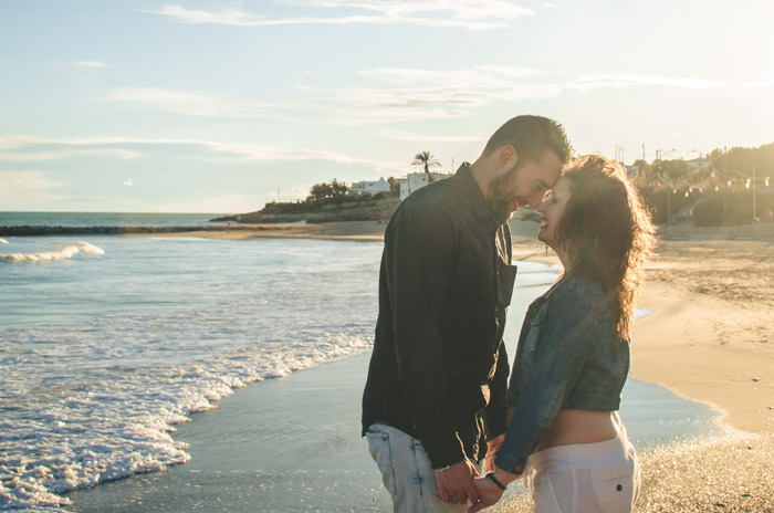 una pareja cerca la una de la otra cogida de la mano en la orilla con la playa y el mar a su alrededor