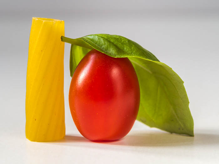 Foto de comida minimalista cerca de un trozo de pasta rigatoni, un tomate ciruela y una hoja de albahaca 