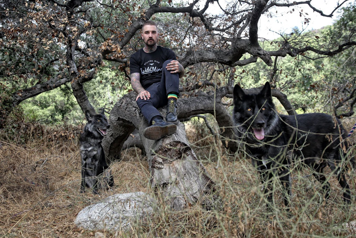 Shot of a man sitting on a tree with a black dog below in dry grass