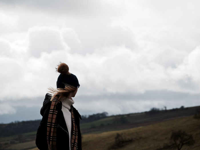 Girl stood in white turtle neck, hat and scarf looking up a hillside.