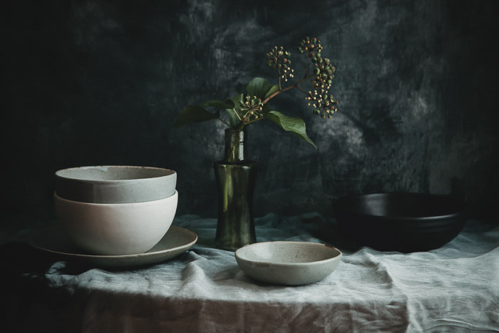Still life photography of pottery bowls and a flower in a vase on a table, with dark background. 