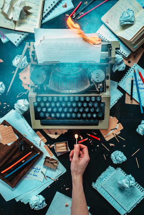 Overhead-Aufnahme einer Schreibmaschine und unordentlicher Utensilien auf dunklem Hintergrund, eine ausgestreckte Hand hält ein brennendes Streichholz - Ideen für Stilllebenfotografie.Stillleben-Fotografie-Ideen