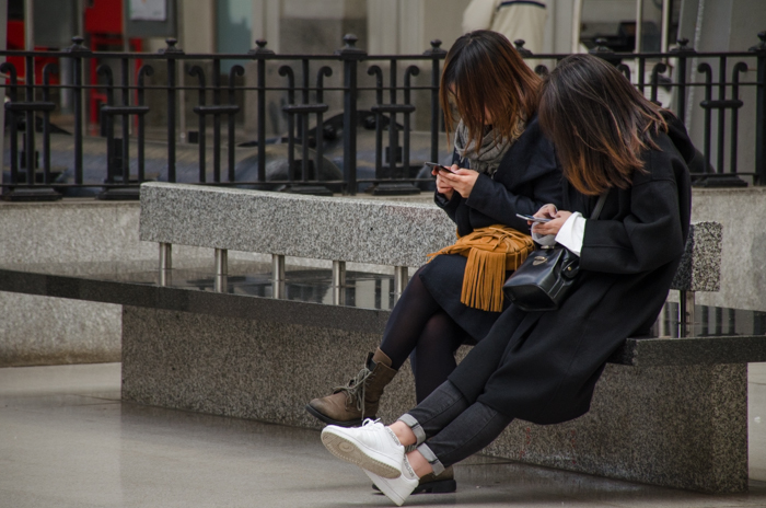Stadsfoto van twee meisjes die op een bankje zitten en een smartphone gebruiken, toont midden- en donkere tinten in afbeeldingen