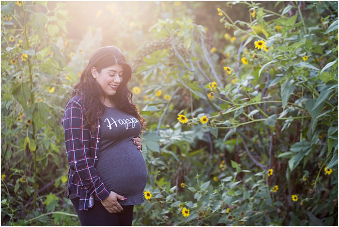 Bidikan fotografi bersalin yang indah dari seorang wanita hamil yang mengenakan kemeja dengan tulisan Happy di atasnya, menggendong bayinya berdiri di taman dengan sinar matahari keemasan di belakangnya