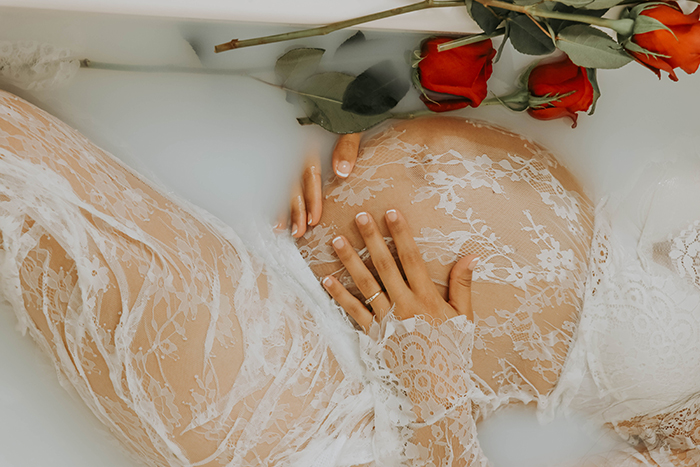 Uma sessão fotográfica de banho de leite em close-up de uma modelo de mulher grávida usando renda branca e segurando a barriga, cercada por flores