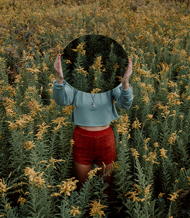 Surreal mirror photography shot of a female model in an overgrown field, holding a mirror over her ace which reflects more overgrown foliage