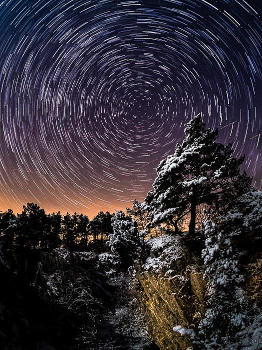 A lone pine tree on a cliff under a classic star trail centred on Polaris. 