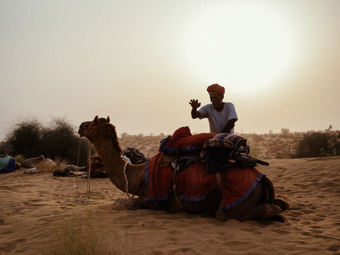 een portret van een man die bij een kameel in de woestijn staat, met een prachtige lensflare achter hem