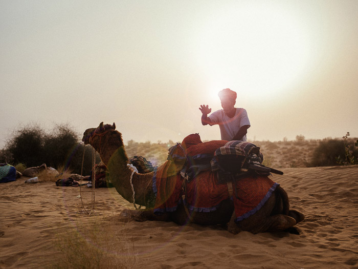een portret van een man die bij een kameel in de woestijn staat, met een prachtig lensflare-effect achter hem