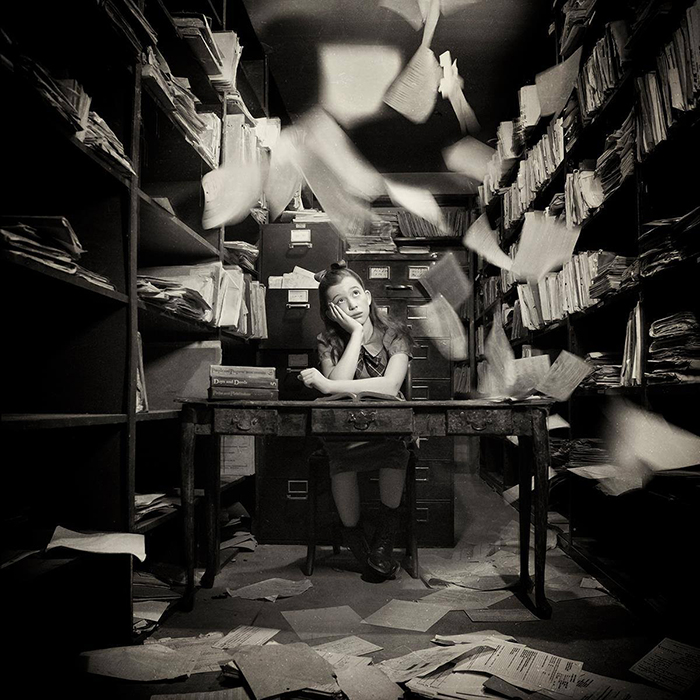 Conceptual black and white portrait of a young girl sitting in an office with papers flying around by fine art photographer Carolyn Hampton
