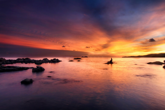 Photo of a waterscape with a fisherman at sunset in purple, orange, and yellow colours
