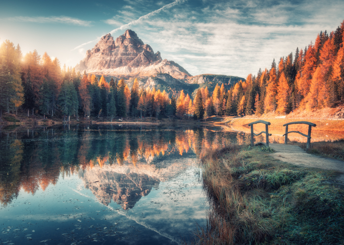Lago com reflexo nas montanhas ao nascer do sol no outono nas Dolomitas, Itália. Paisagem com o Lago Antorno, pequena ponte de madeira, árvores com folhas de laranja, pedras altas, céu azul no outono. Floresta colorida