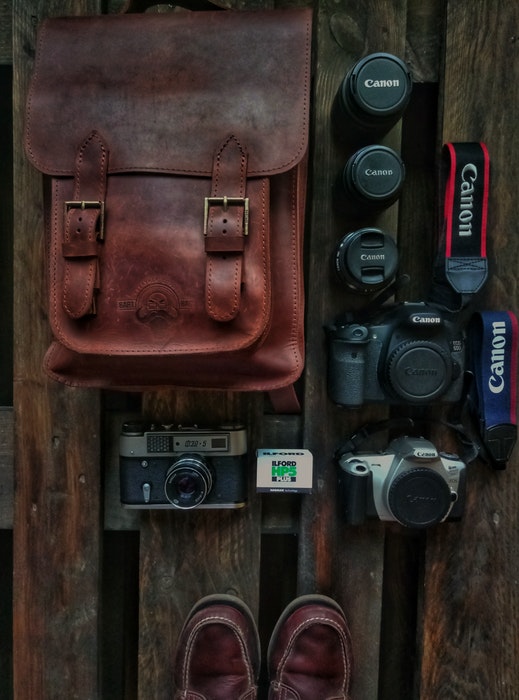 Uma foto aérea de uma bolsa de couro para câmera e equipamentos espalhados por uma superfície de madeira.