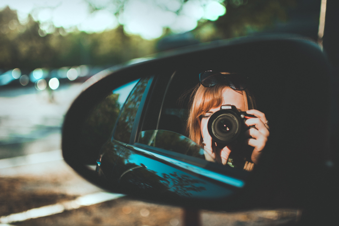 Um reflexo no espelho lateral de uma mulher com uma câmera DSLR no banco de trás de um carro