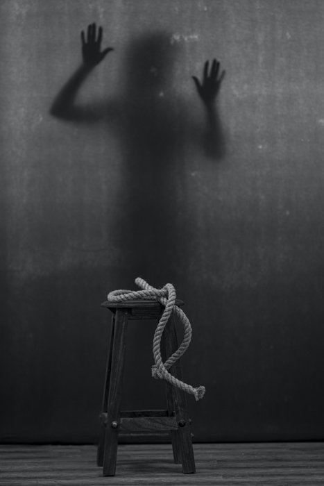 imagem em preto e branco de uma corda em um banquinho e uma figura sombria no fundo 